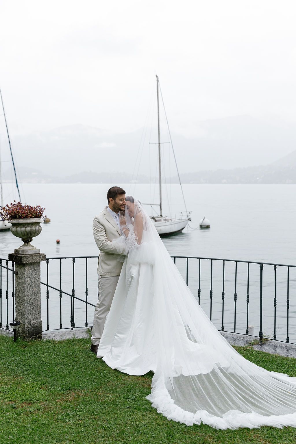 A wedding couple poses along a lakeside railing with mountains and boats in the background, showcasing a flowing white dress in Lake Como.