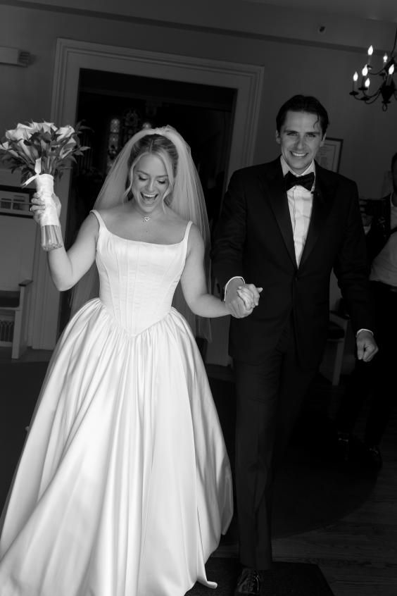 Black and white wedding photo showing joyful couple walking together, bride in classic gown holding bouquet.