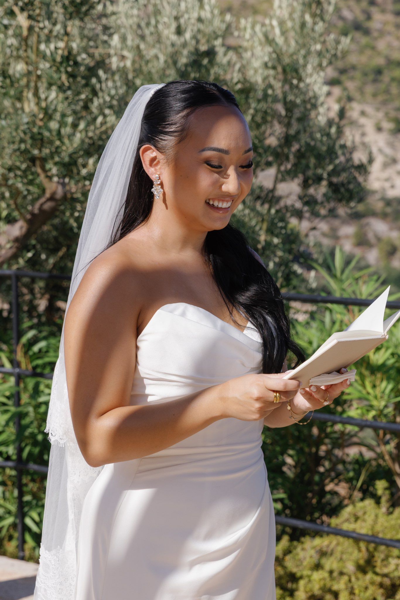 Smiling bride in white strapless gown and veil reading wedding vows from a book outdoors.