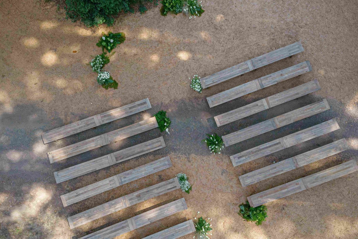 A series of wooden benches arranged in a row under dappled sunlight and tree shadows on a dirt ground.