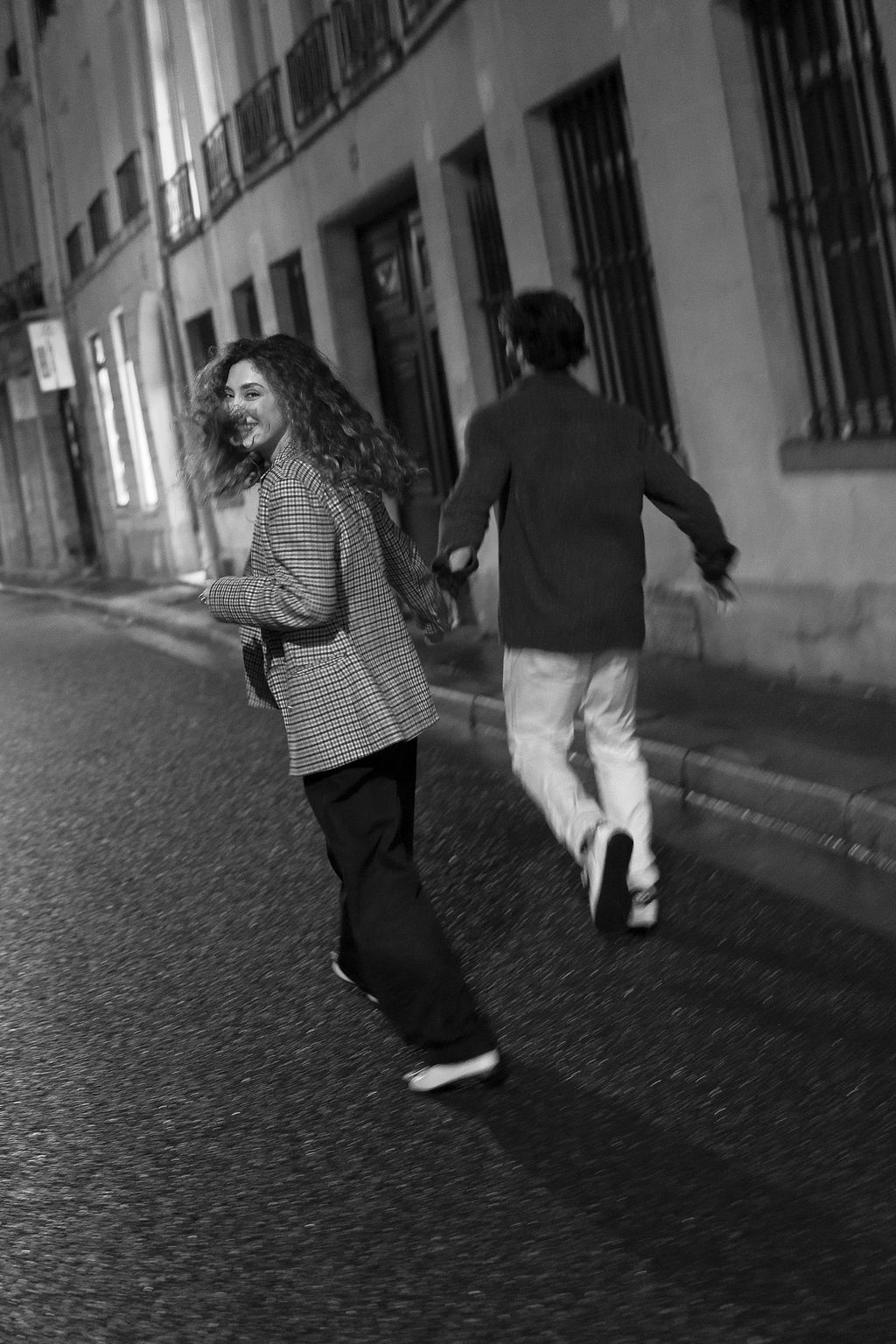 A black and white street photograph showing two people walking and laughing together on a cobblestone road in Paris.