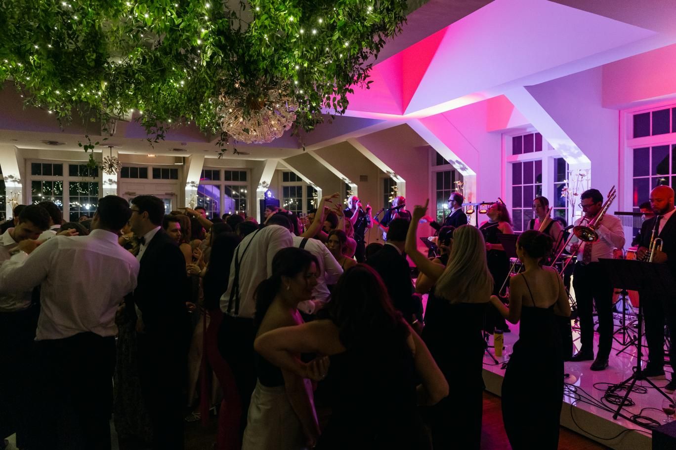 Crowded dance floor at an evening reception with pink and purple uplighting illuminating the venue's white columns and ceiling.