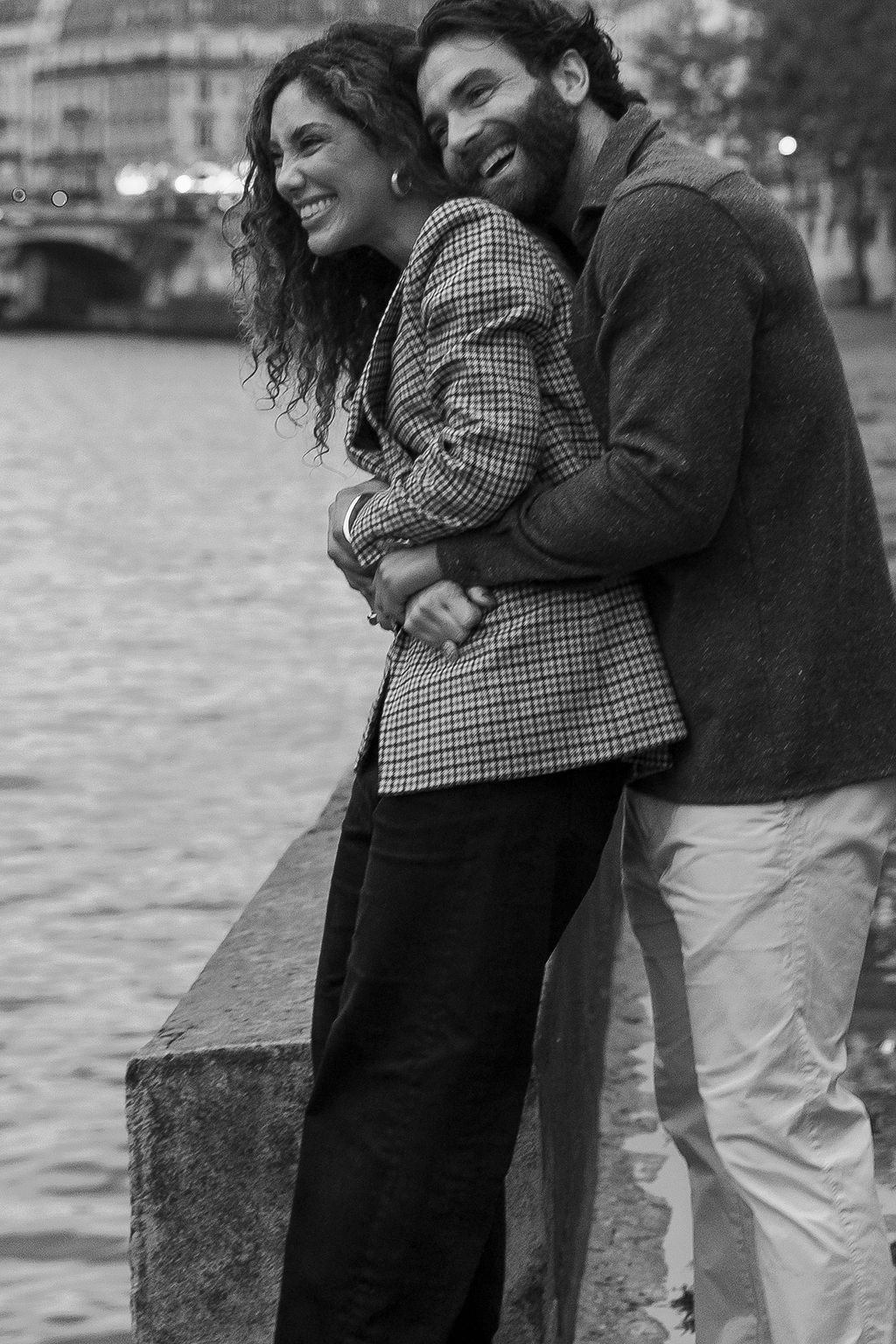 A couple shares a romantic moment while standing by the water's edge in a black and white photograph in Paris, France.