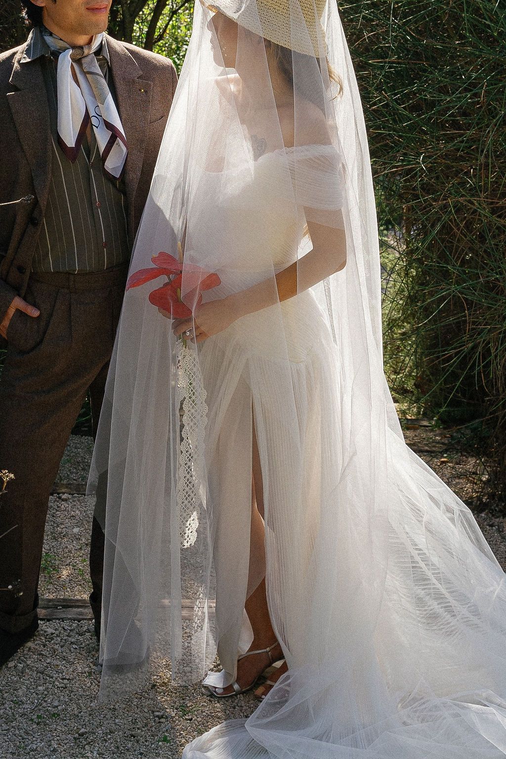 Elegant bride in flowing white veil and dress stands beside figure in brown suit outdoors in Tuscany, Italy.