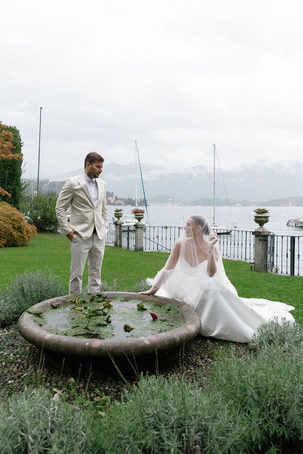 Wedding couple interacts playfully around a stone fountain in a lakeside garden with mountains in the background in Lake Como.