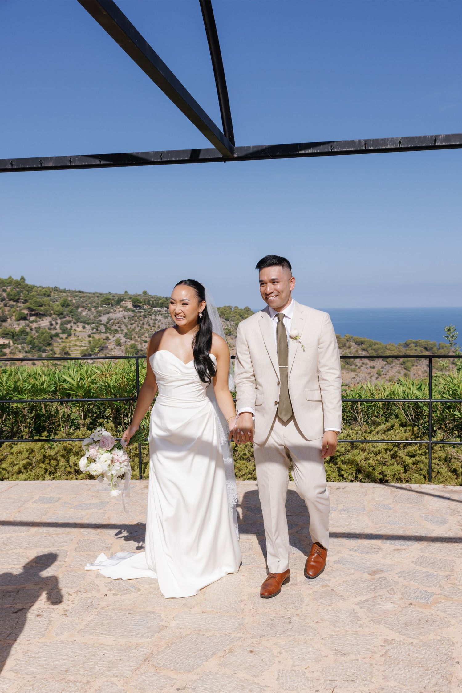 Newlywed couple holds hands smiling on stone terrace with lush green hills and blue ocean backdrop.