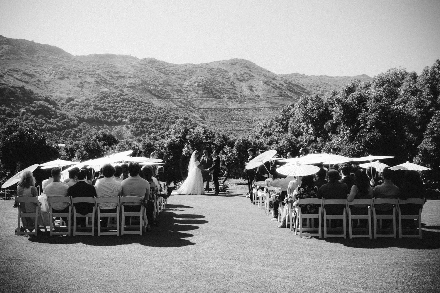 Black and white outdoor wedding ceremony with guests seated in white chairs facing mountain landscape backdrop.