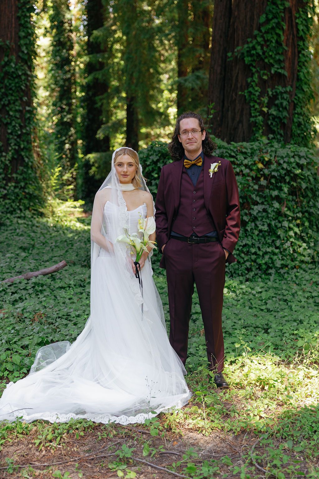 Wedding couple posing in a lush forest setting with ivy-covered trees and dappled sunlight.