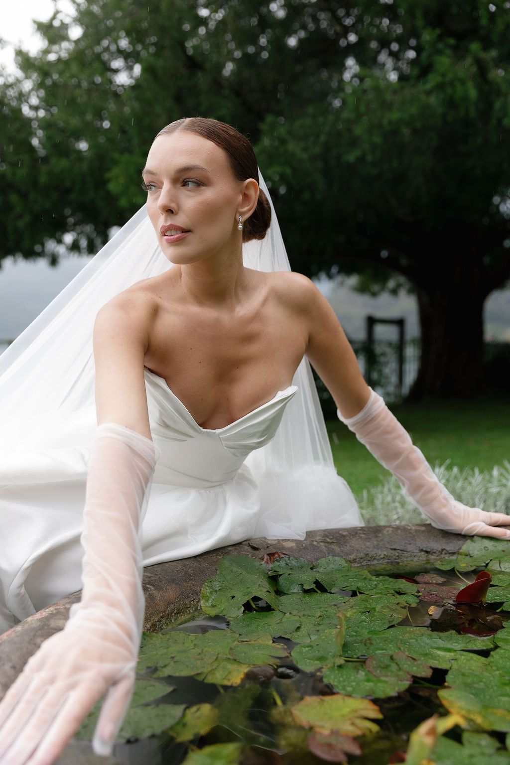 Bride in strapless gown and sheer gloves poses by lily pads in garden setting in Lake Como.