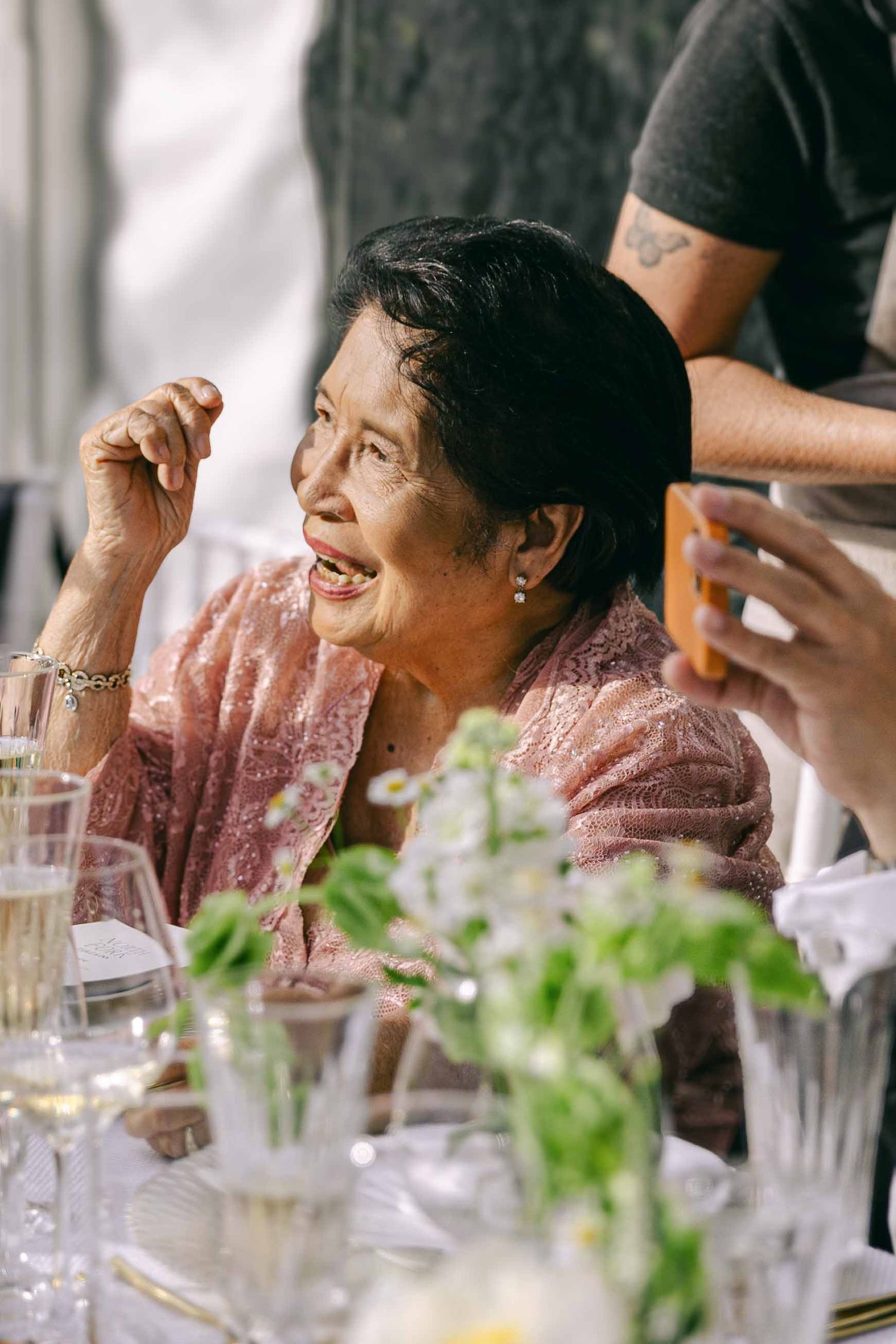 Elderly woman in pink lace laughing joyfully at an elegant outdoor dining table with floral arrangements.