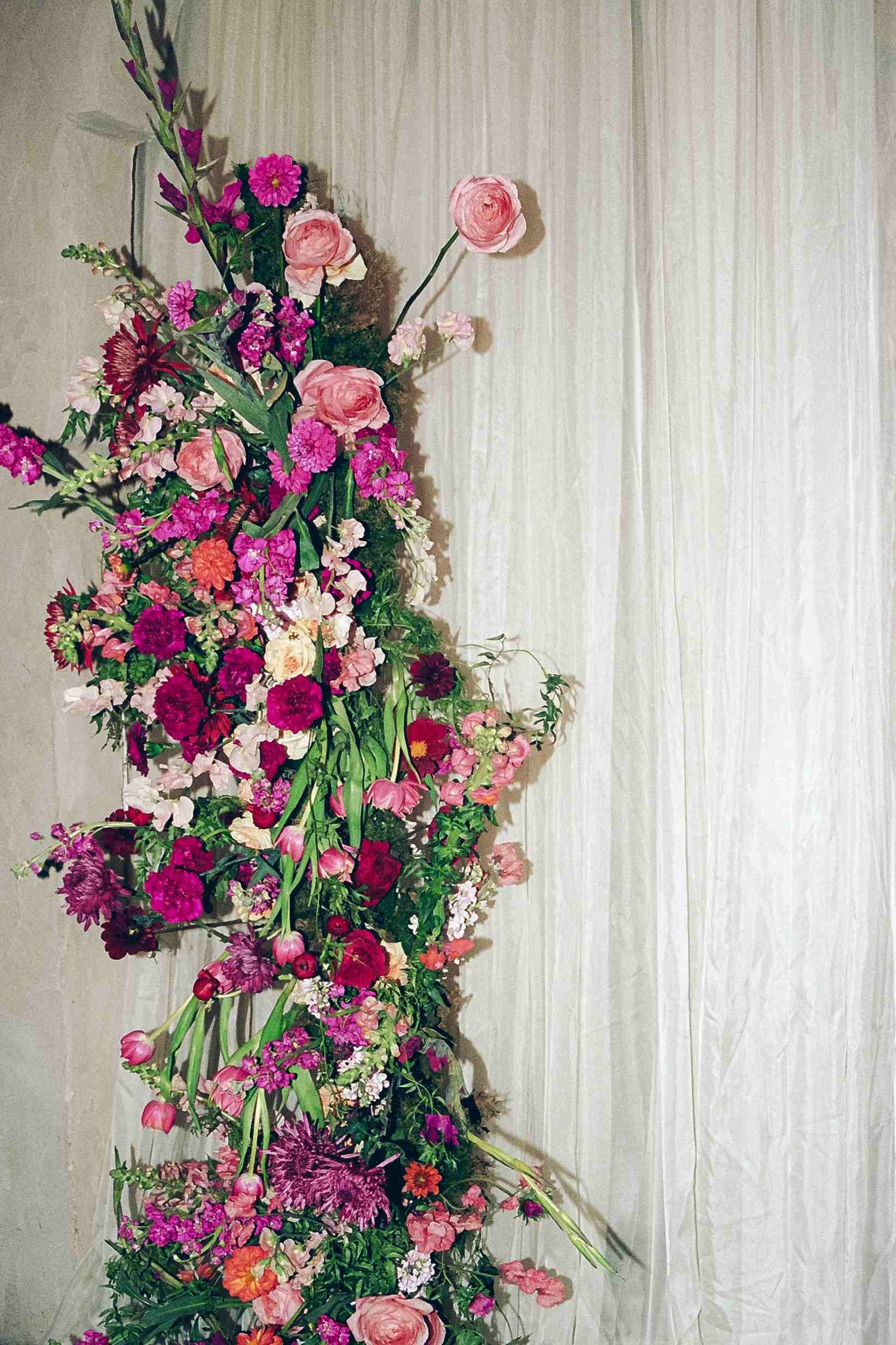 A vertical floral arrangement with pink roses and purple blooms cascades against a light colored wall.