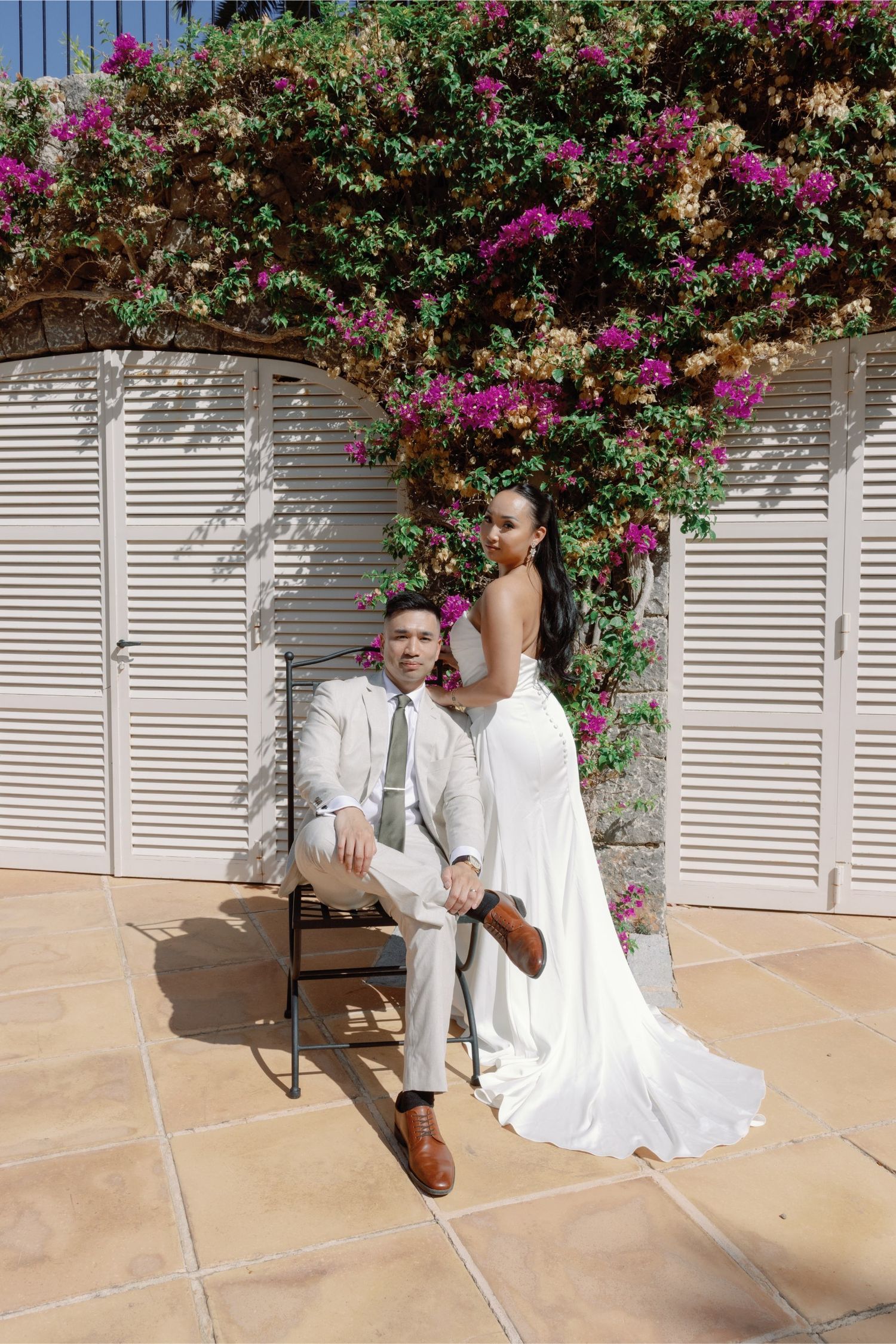 Bride in white gown stands behind groom in gray suit seated on chair under blooming bougainvillea on sunny patio.