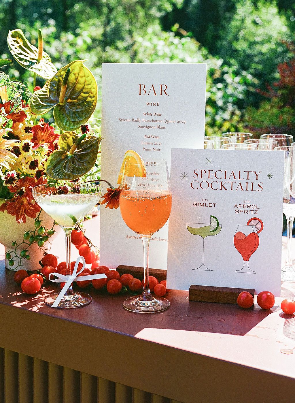 A wedding bar menu display with specialty cocktail cards and floral decorations on a brown table.