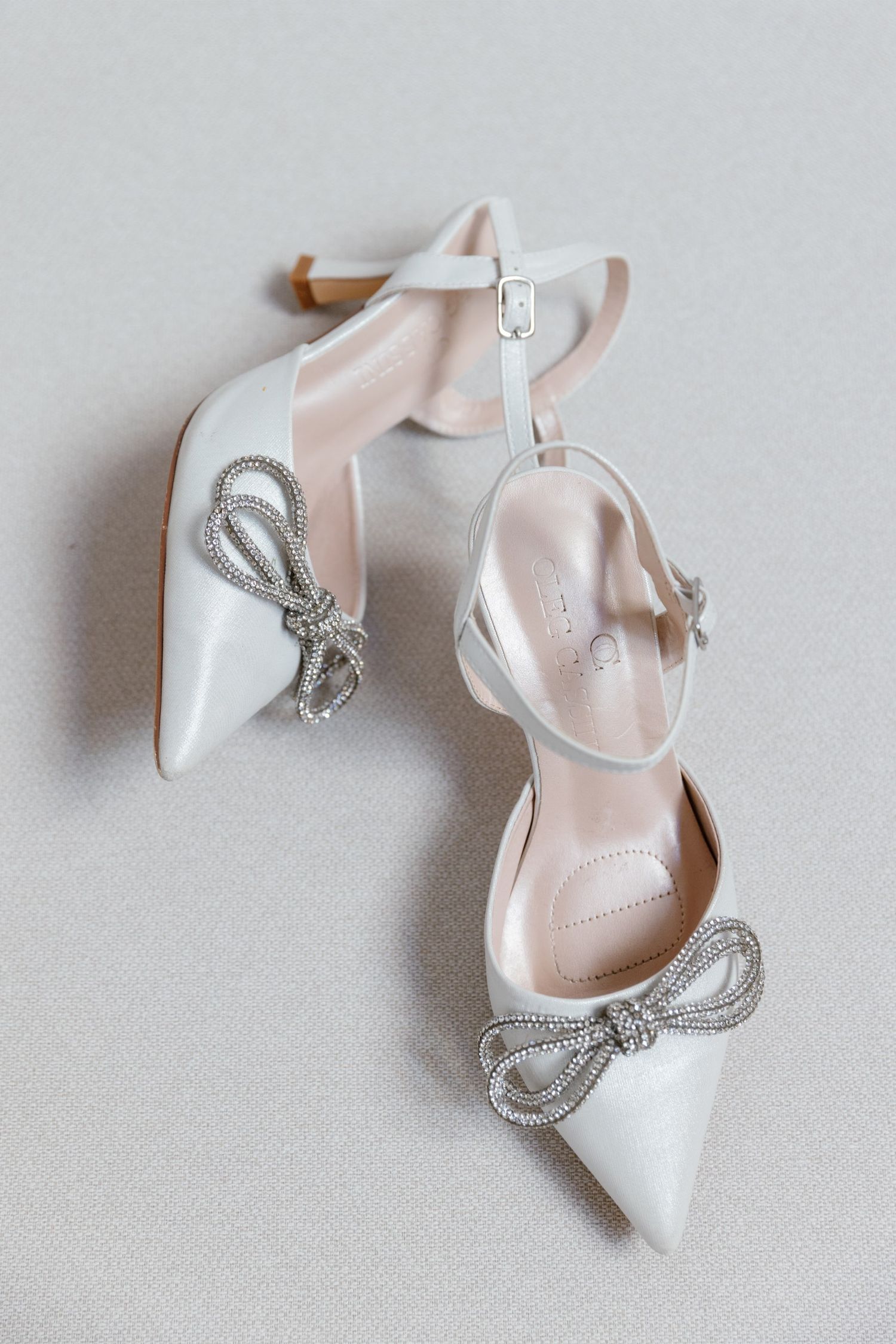 White satin bridal heels with crystal bow detail and ankle strap on a neutral background.