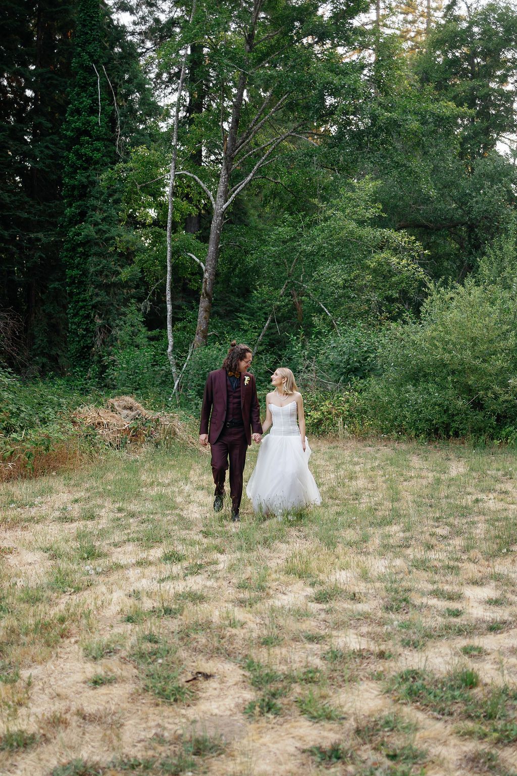 A couple walking together through a grassy field surrounded by trees on a sunny day.