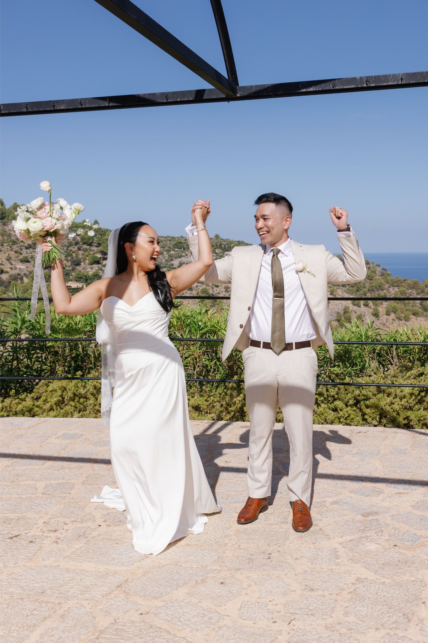 Joyful newlywed couple celebrating after wedding ceremony outdoors with ocean views, bride in white gown holding bouquet.