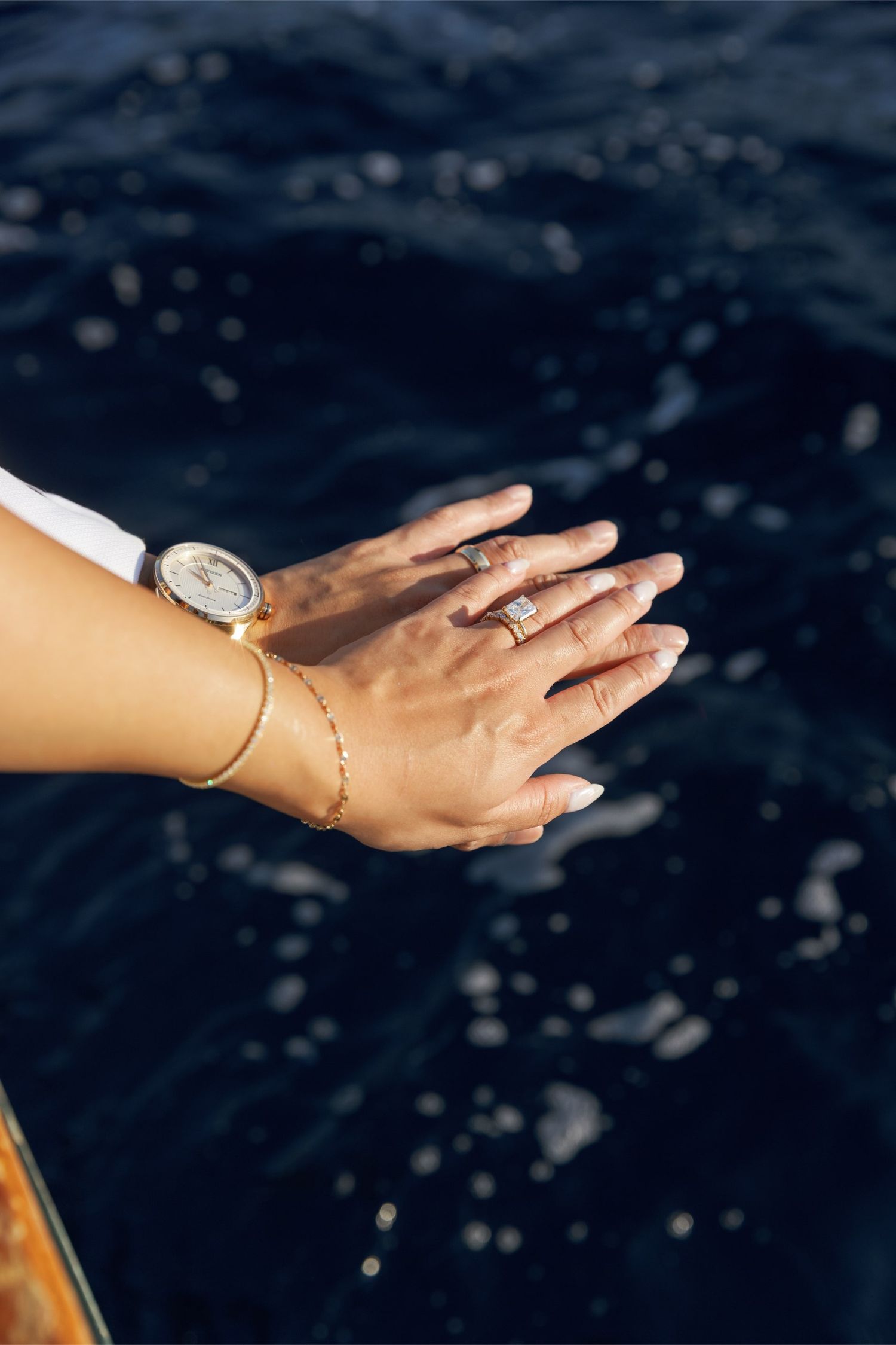 Two hands adorned with a diamond ring, gold watch, and delicate bracelet posed over sparkling blue ocean water.