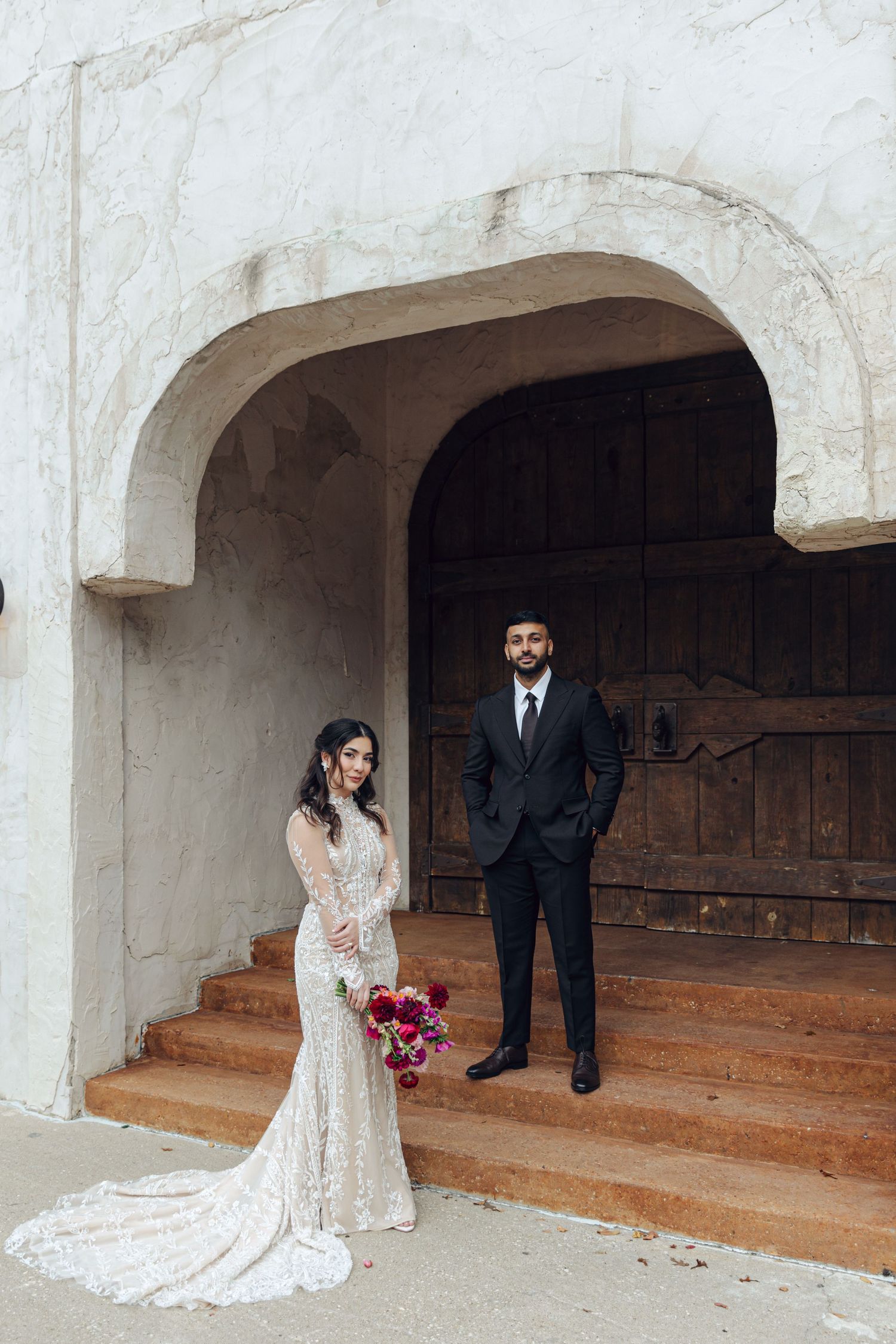 A bride in a lace wedding dress with a bouquet stands with a groom in a black suit under a white archway entrance.
