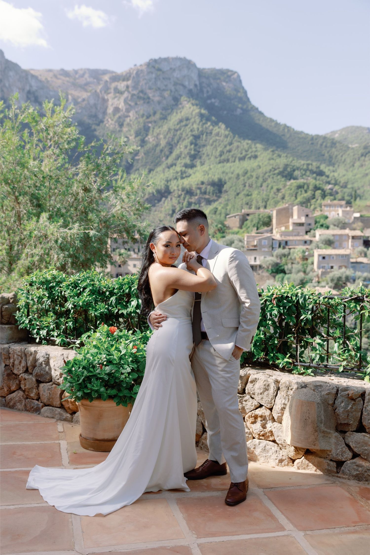 Bride and groom embrace on a terrace with lush green mountains and a stone village in the background.