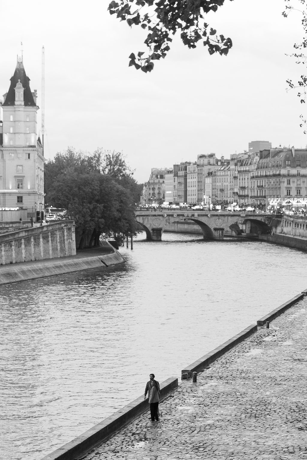 Black and white photograph of a lone figure walking along the Seine River in Paris with historic buildings and bridges in view.