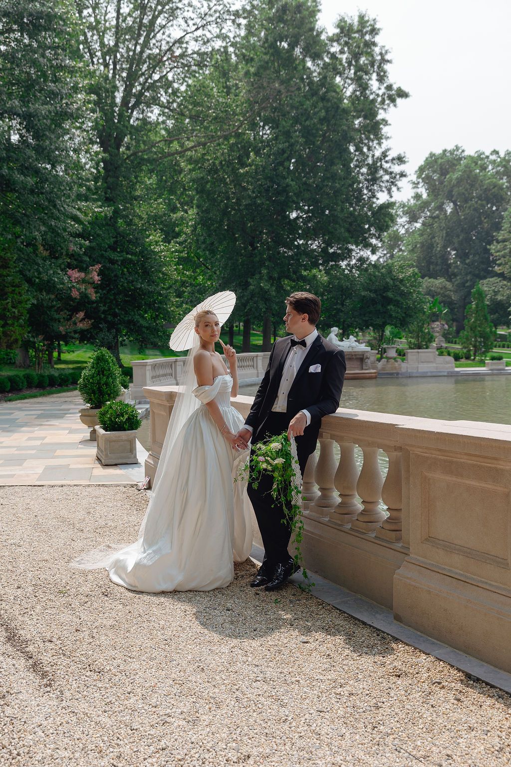 Wedding couple sitting on stone balustrade with lush green trees in background.