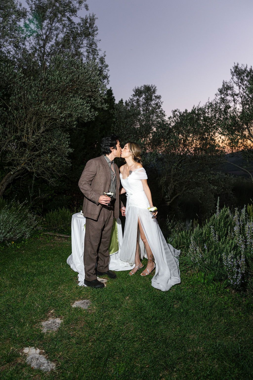 A couple in formal attire shares a romantic moment in a garden with trees silhouetted against the evening sky in Tuscany, Italy.