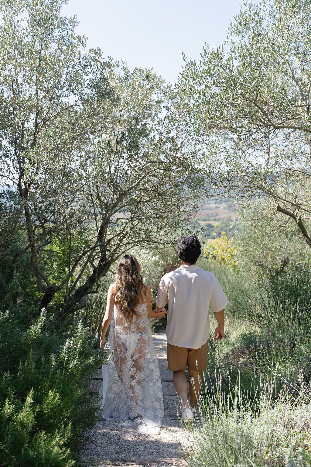 A couple walks hand in hand down a scenic garden path surrounded by olive trees in the Mediterranean countryside in Tuscany, Italy.