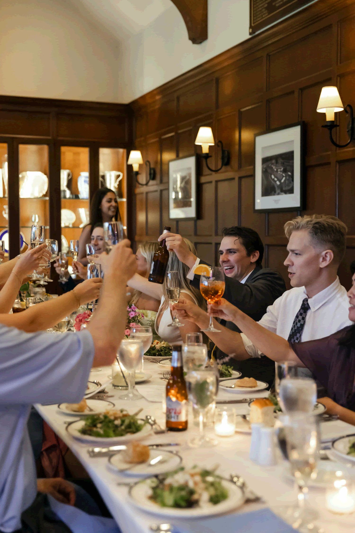 Group of wedding guests raising glasses for a toast during an elegant dinner reception in a wood-paneled dining room.