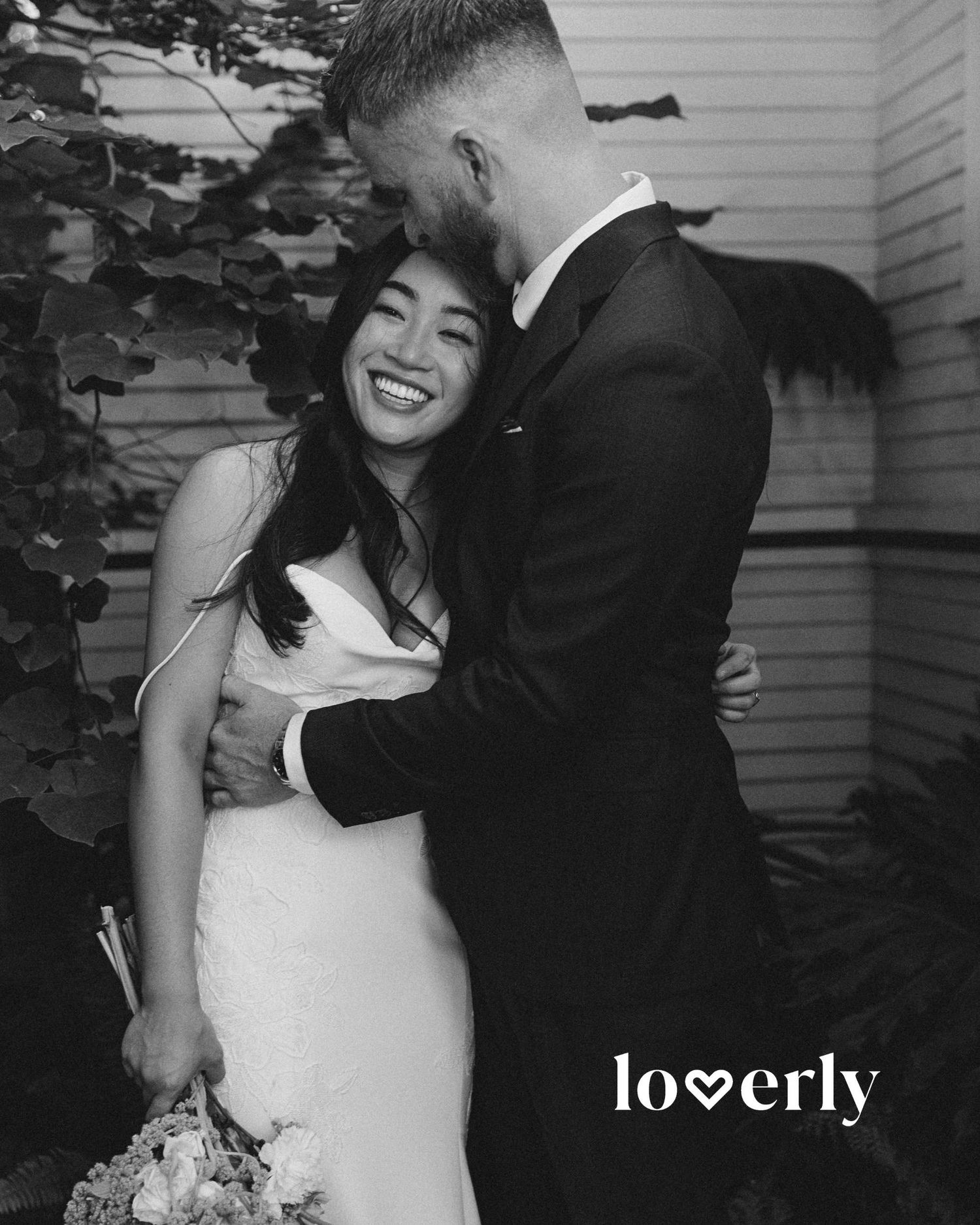 A black and white wedding photo captures an intimate moment between newlyweds sharing a loving embrace outdoors.