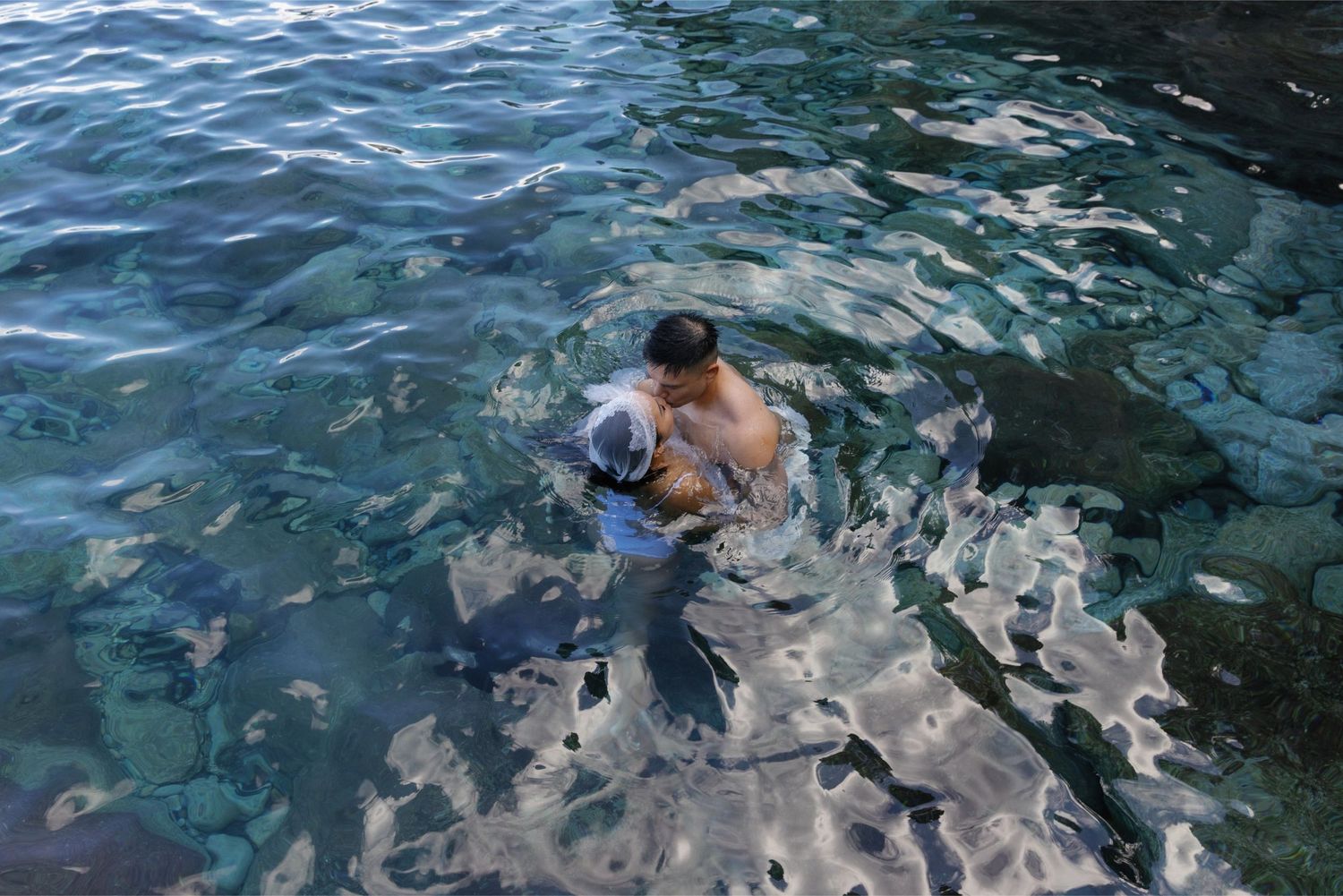Person swimming in clear turquoise ocean water near rocky shoreline, viewed from above.