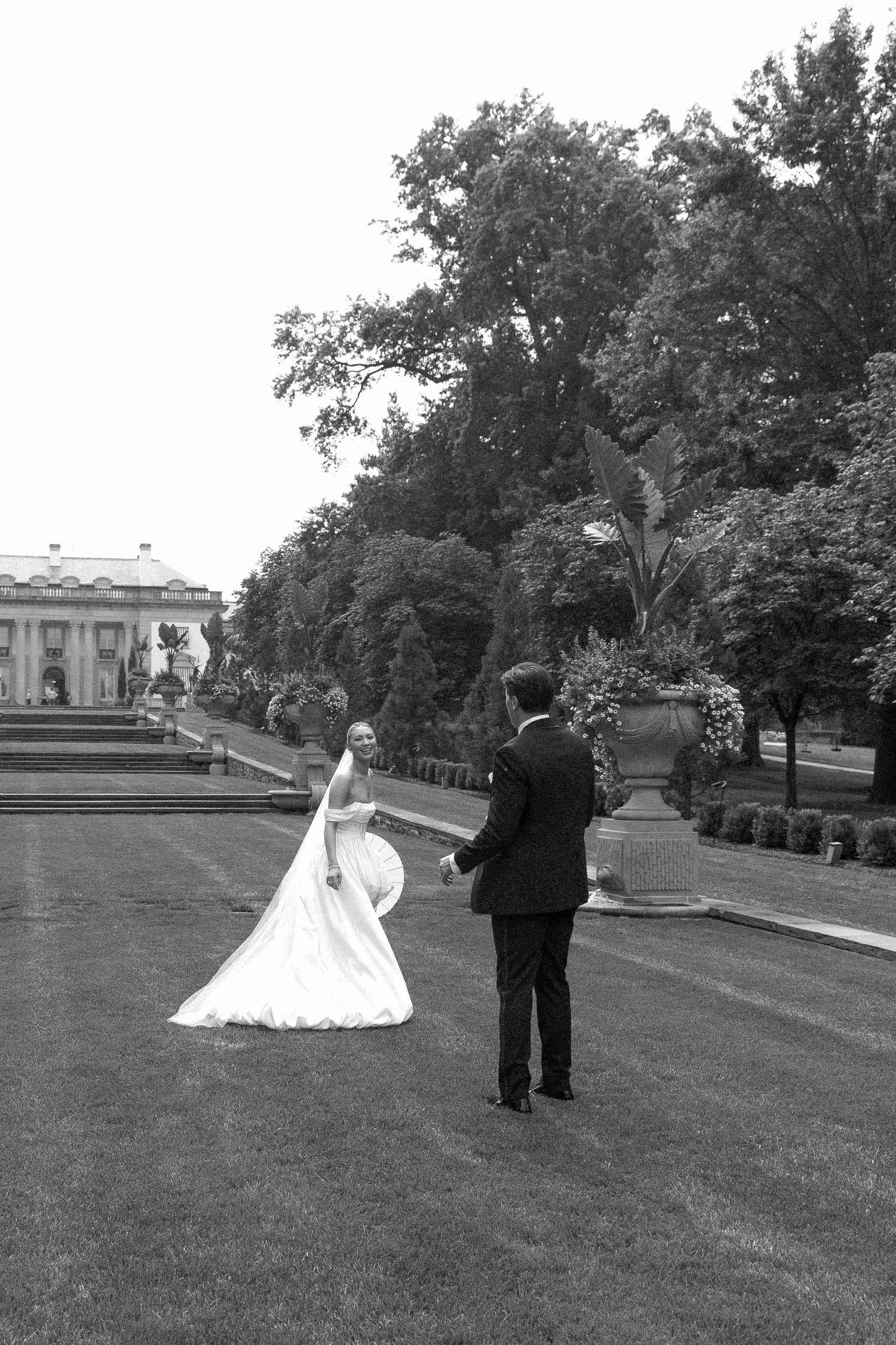 Black and white photo of a bride and groom walking hand in hand across a lush garden lawn at their wedding.