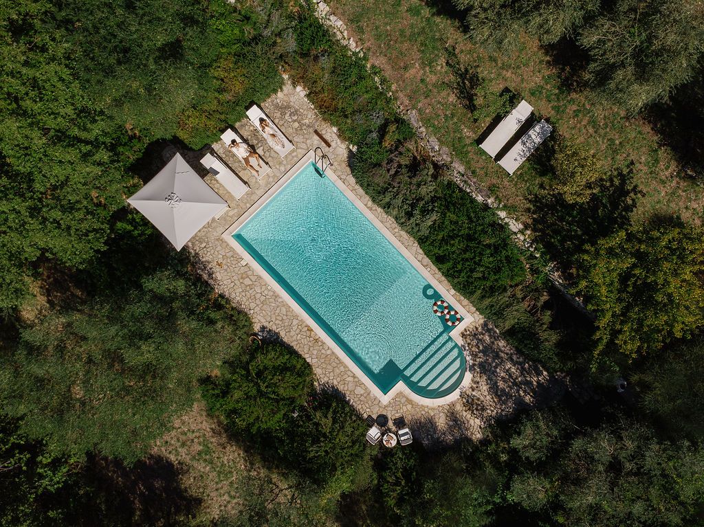 Aerial view of a rectangular swimming pool surrounded by trees and sunbeds in a rustic setting in Tuscany, Italy.