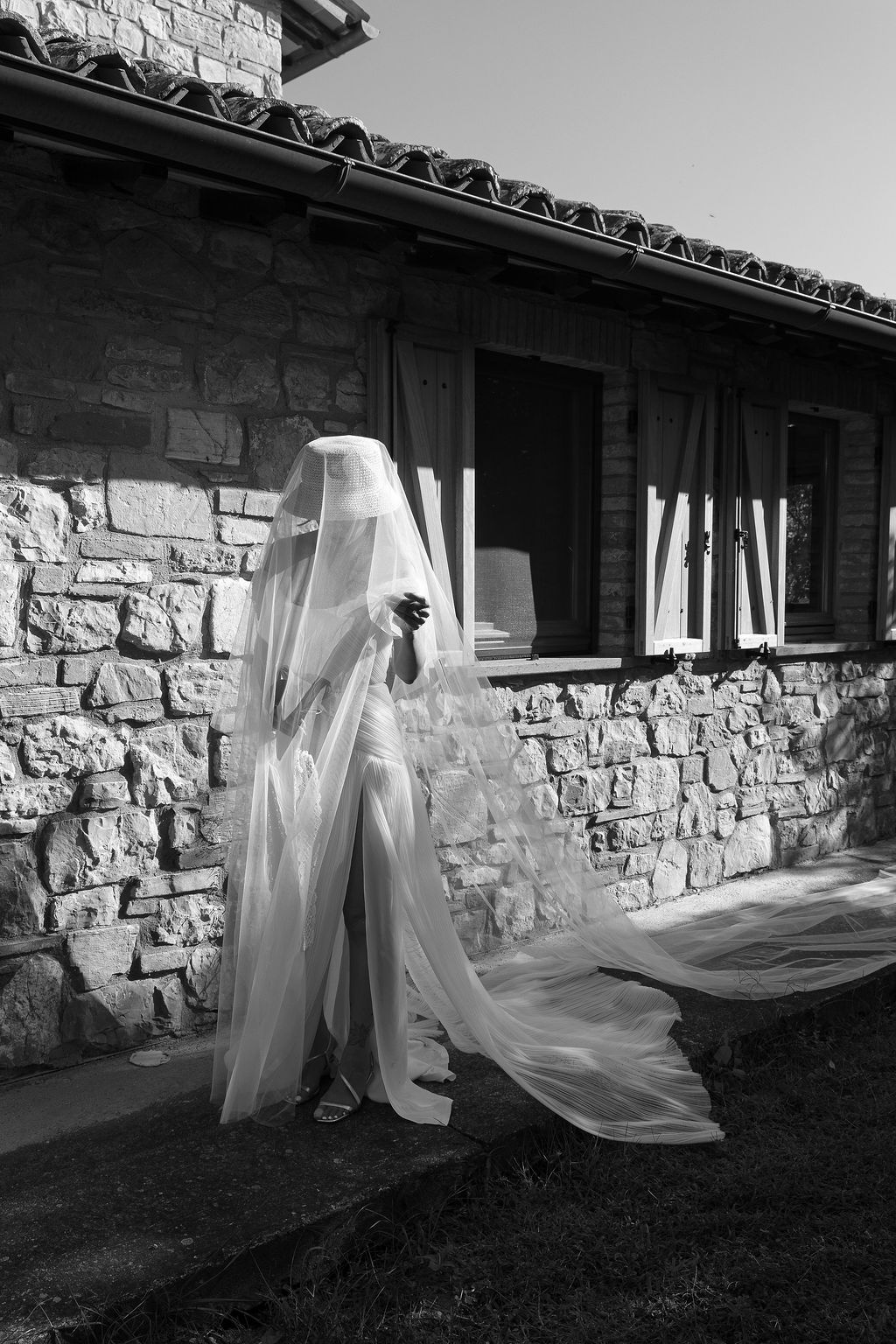 A flowing white wedding dress and veil captured in black and white against a rustic stone wall exterior in Tuscany, Italy.