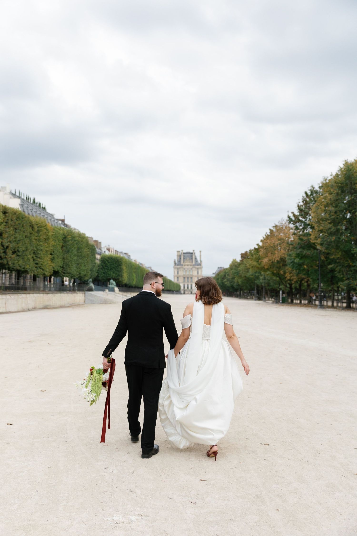 Bride and groom walking hand in hand through Tuileries Garden in Paris, with the Louvre visible in the background.