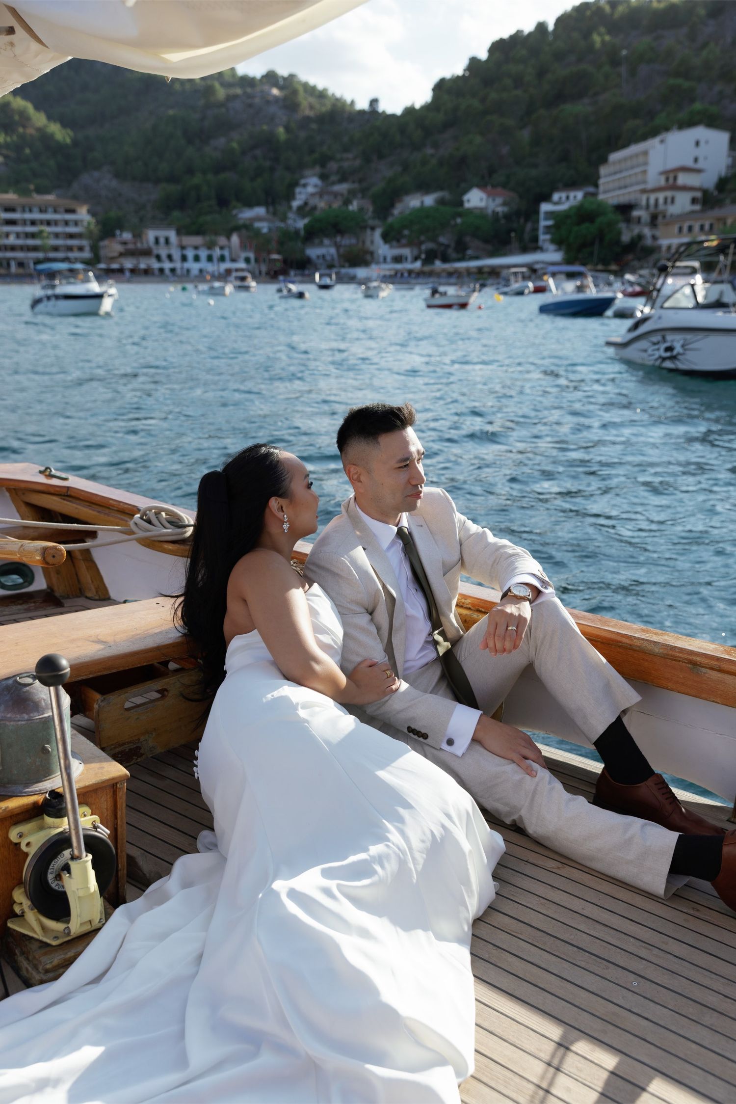Bride and groom sitting together on a wooden boat deck with marina and mountains in the background.