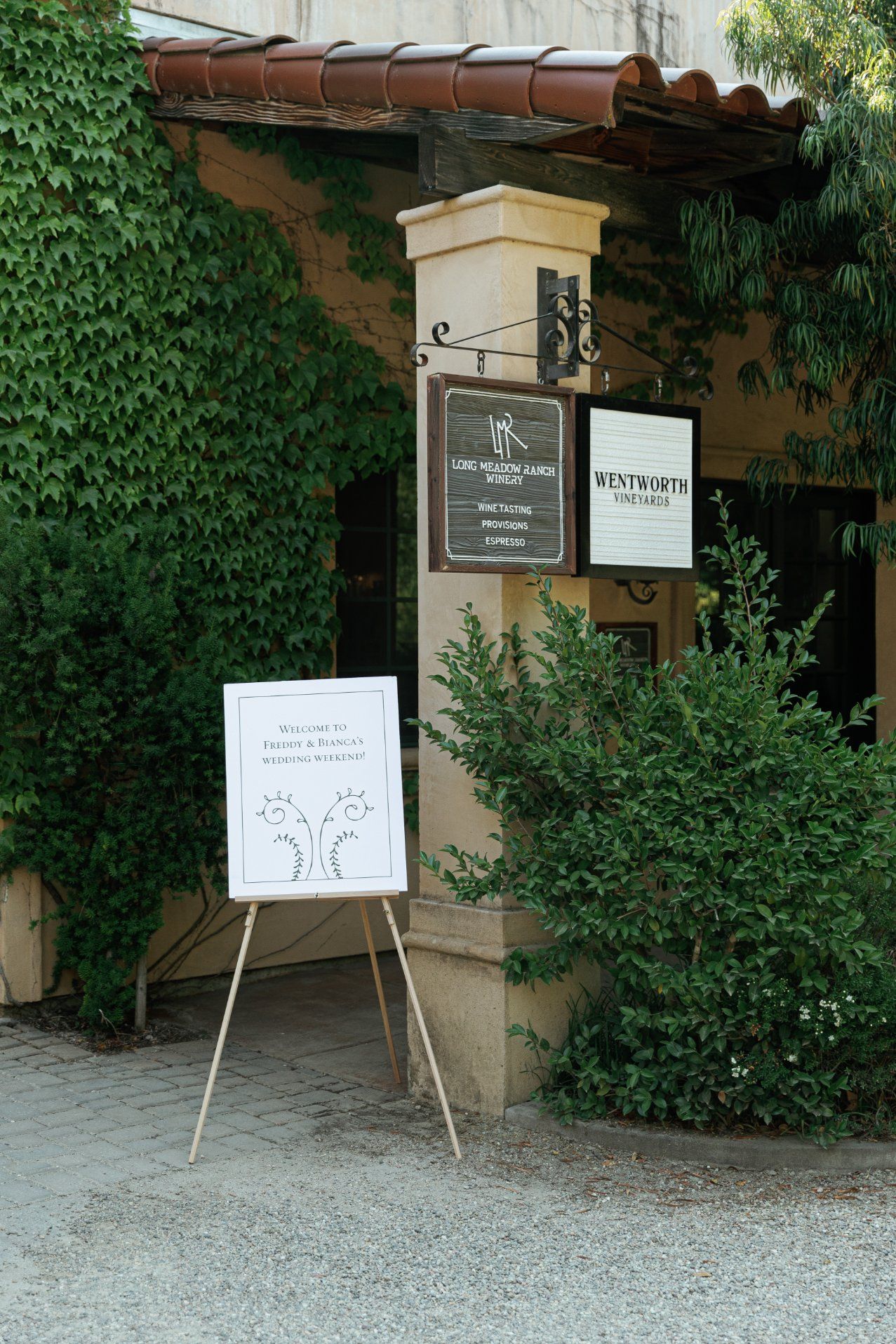 Restaurant entrance with ivy-covered walls, signage, and a whiteboard stand on a gravel pathway.