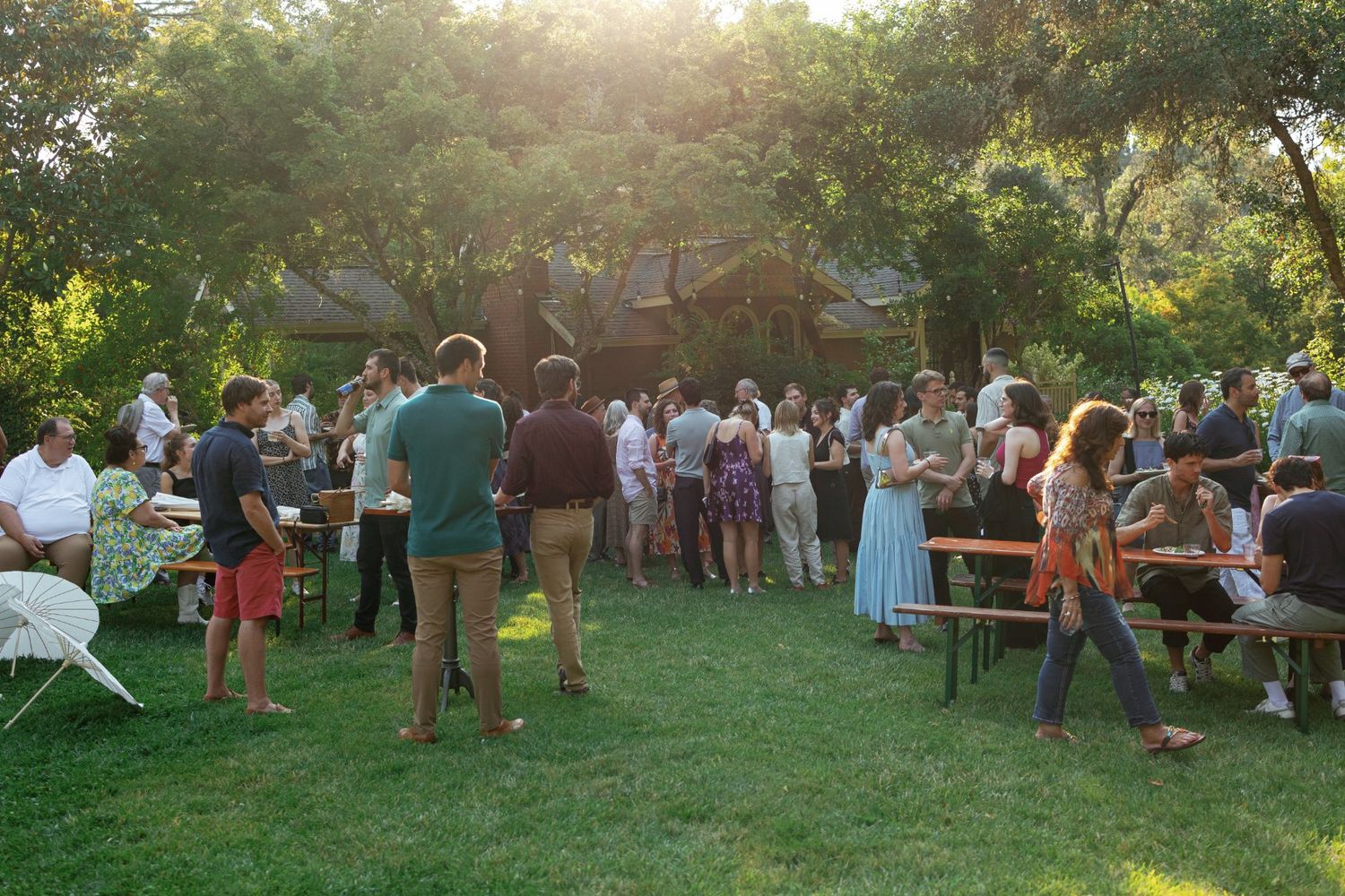 Guests gathered outdoors at sunset for a summer garden party with golden sunlight filtering through the trees.
