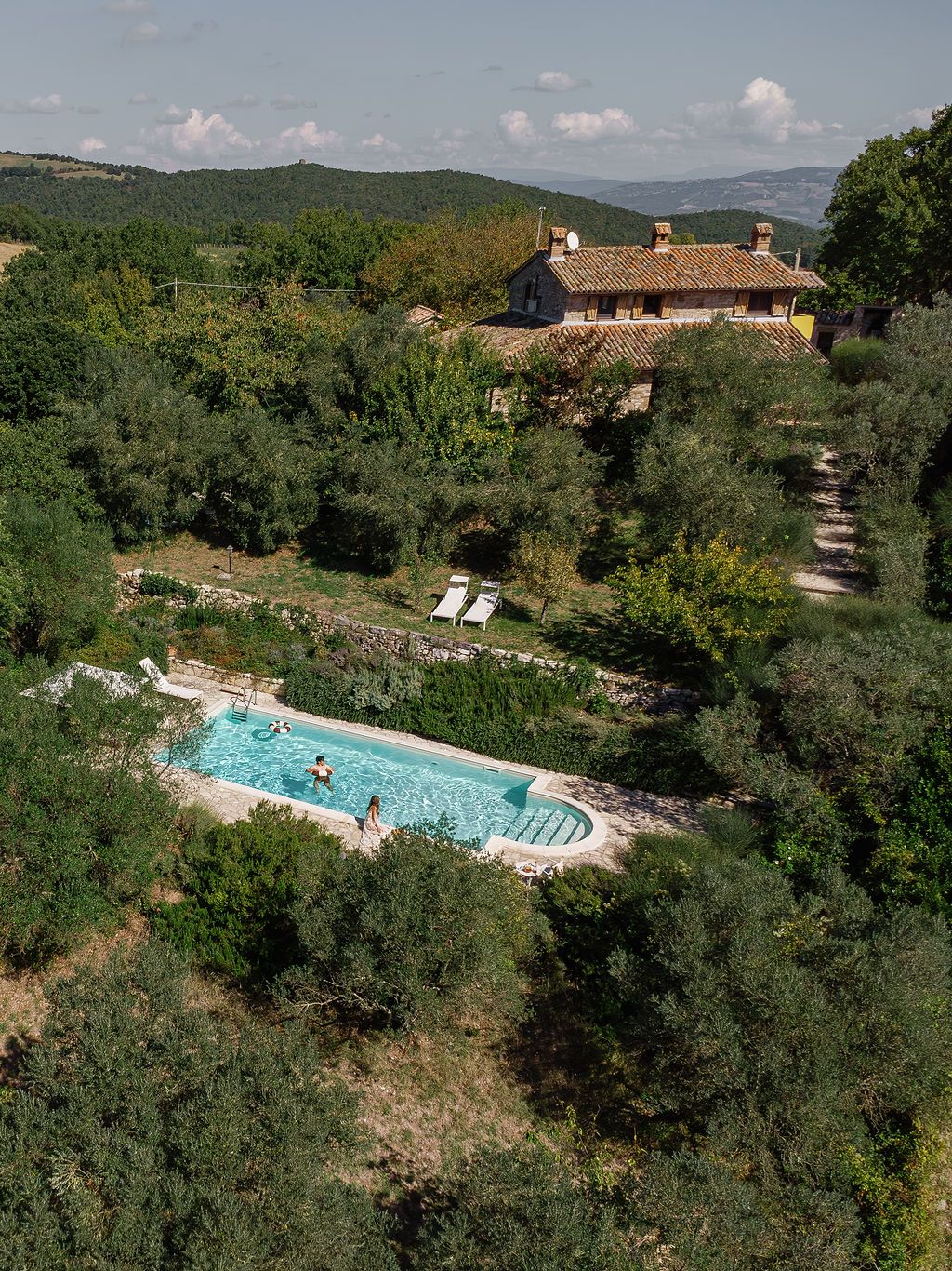 Aerial view of a luxury swimming pool surrounded by lush greenery and rustic Tuscan villas in the countryside in Tuscany, Italy.