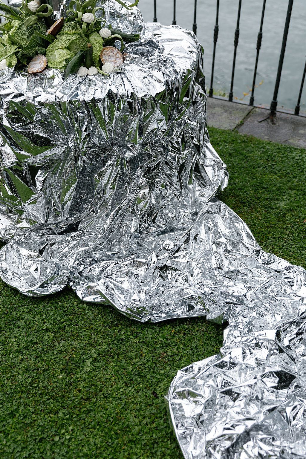 A series of plants wrapped in reflective aluminum foil sit on green grass near a metal fence in Lake Como.