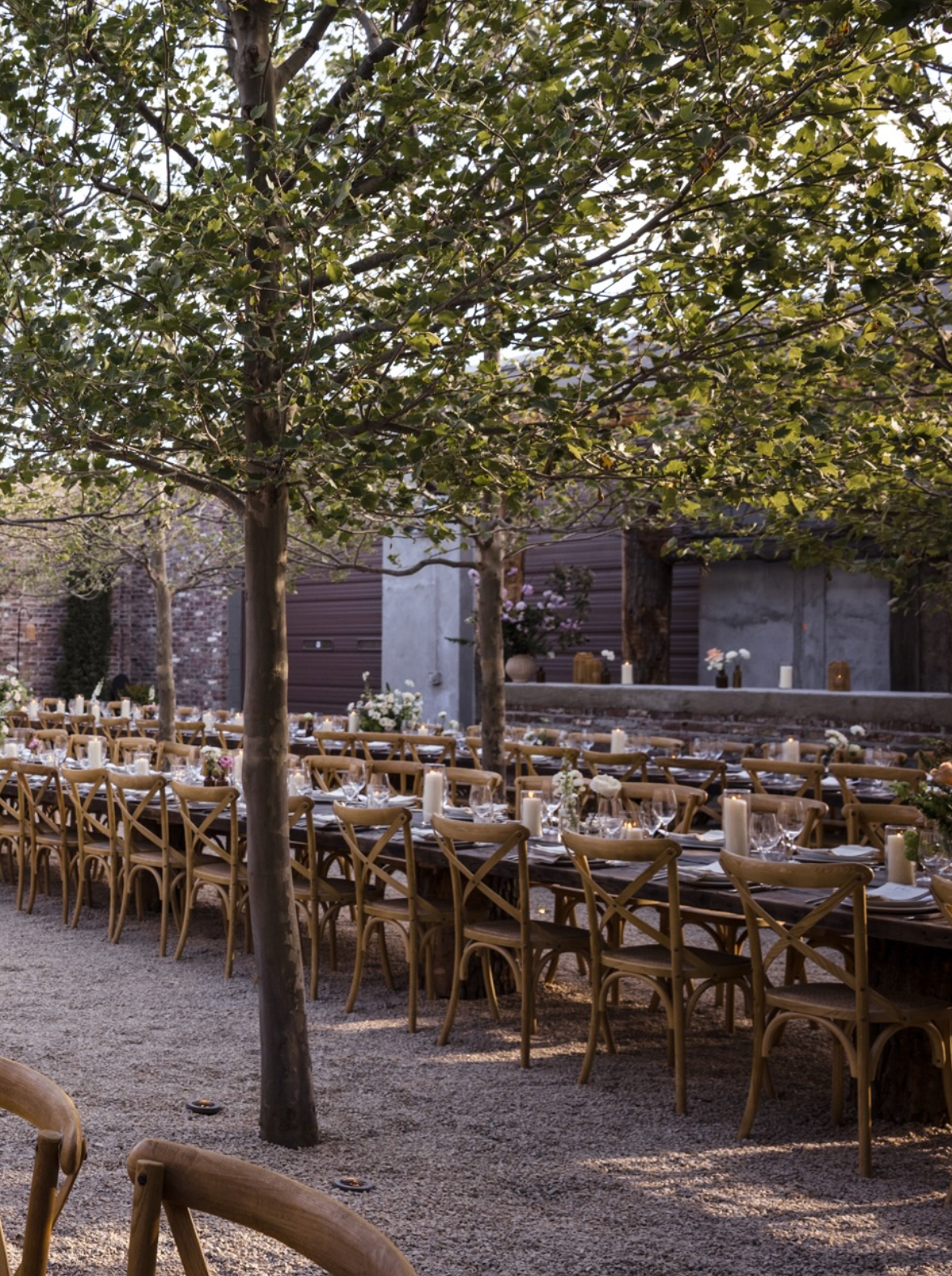 Outdoor wedding reception with long wooden tables, cross-back chairs, gravel ground, and trees in a rustic courtyard setting.