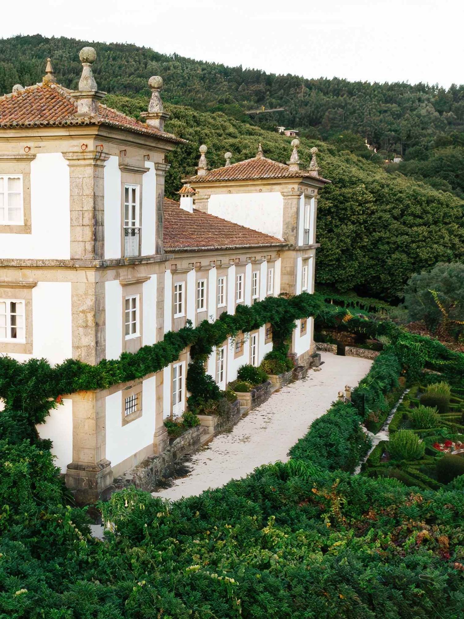 Historic Portuguese manor house with white walls and terracotta roof nestled among lush green gardens on a hillside.