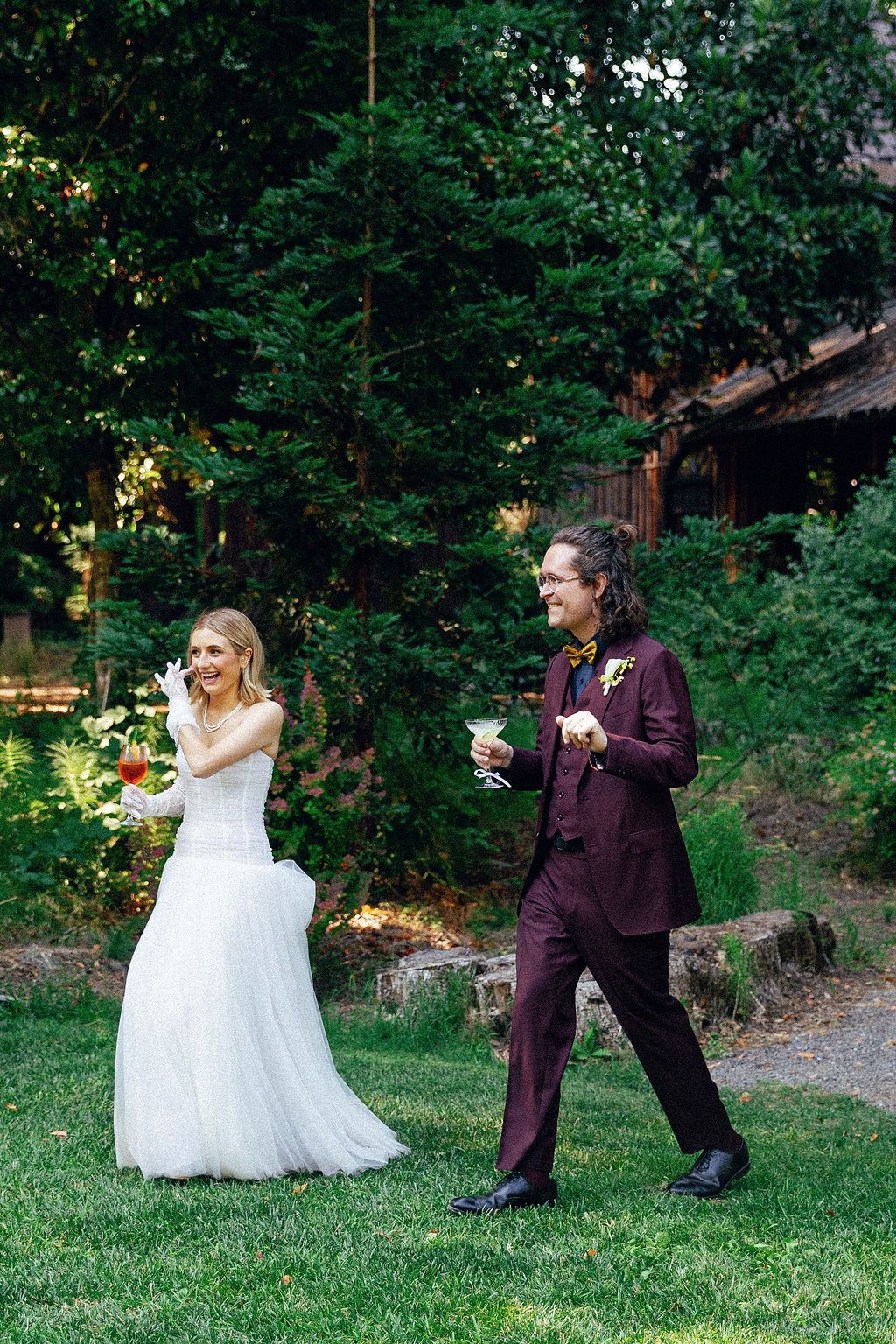 Playful wedding couple poses in a garden setting with burgundy suit and white gown.