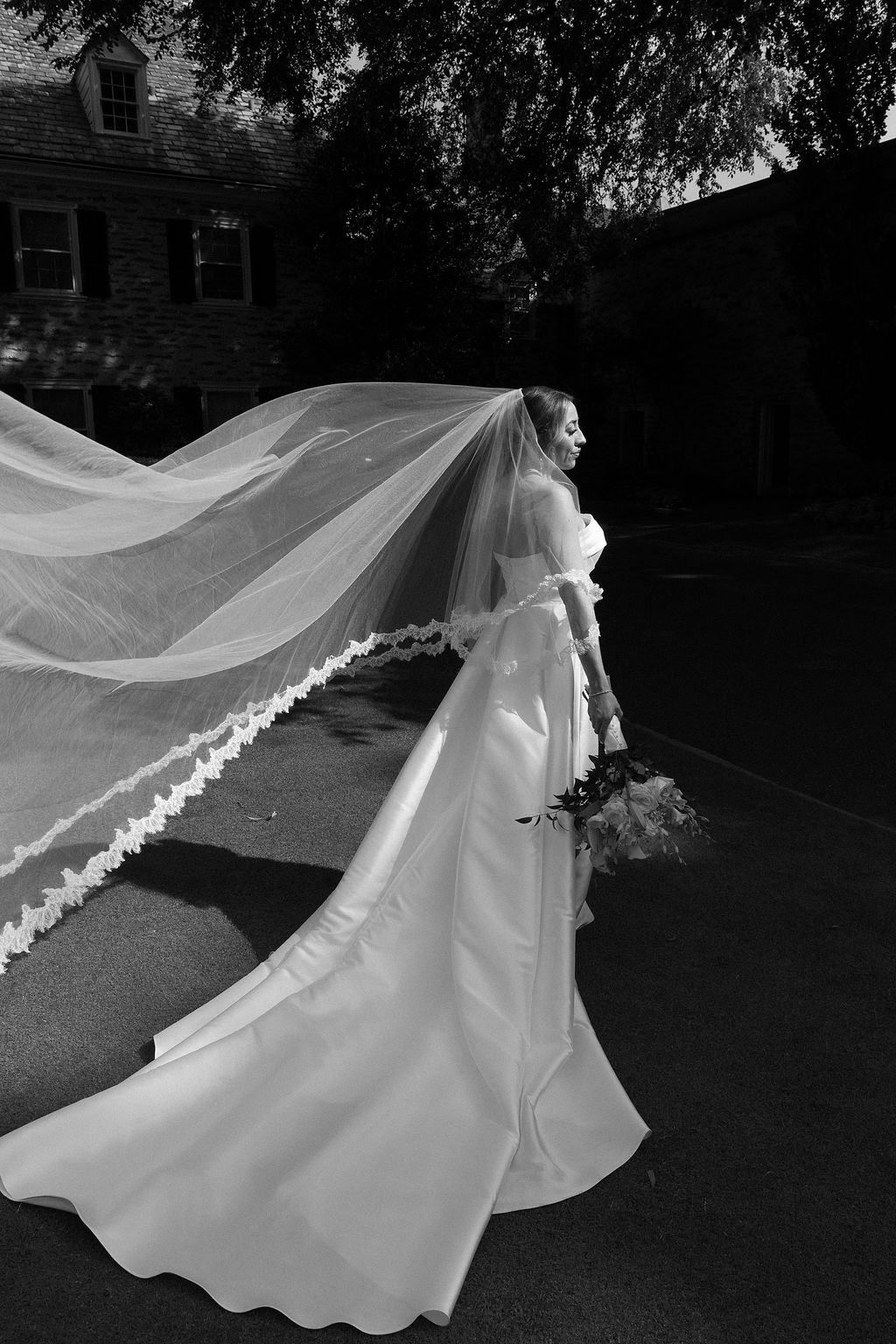 A black and white photograph of a bride in an elegant white gown with a flowing veil captured outdoors.