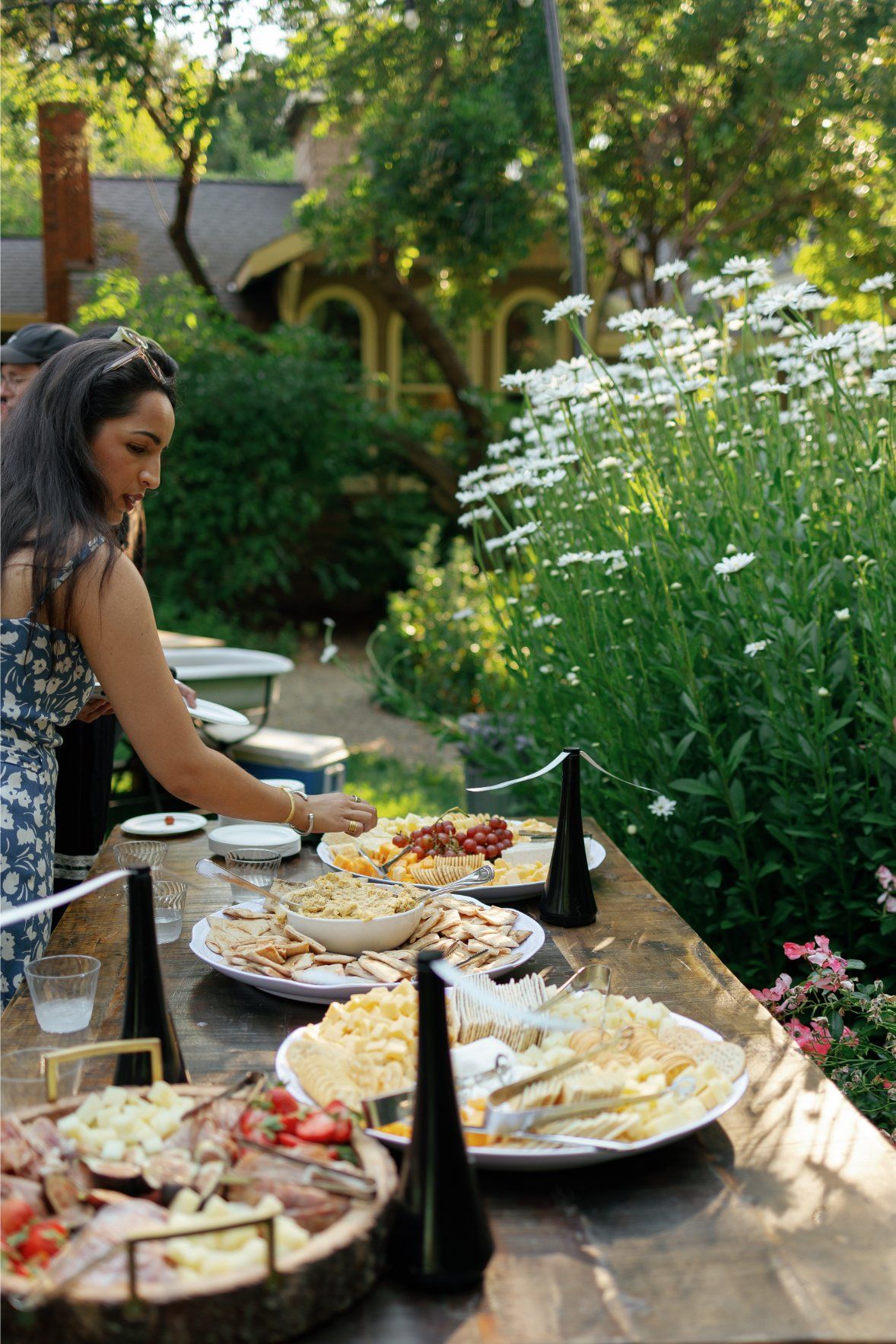 A garden party spread featuring appetizers, cheese boards, and wine set among blooming flowers and greenery.
