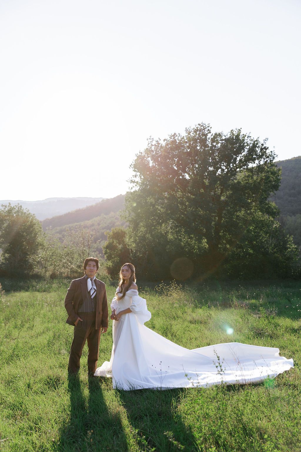 Wedding couple posing in a sunlit meadow with mountains in the background in Tuscany, Italy.