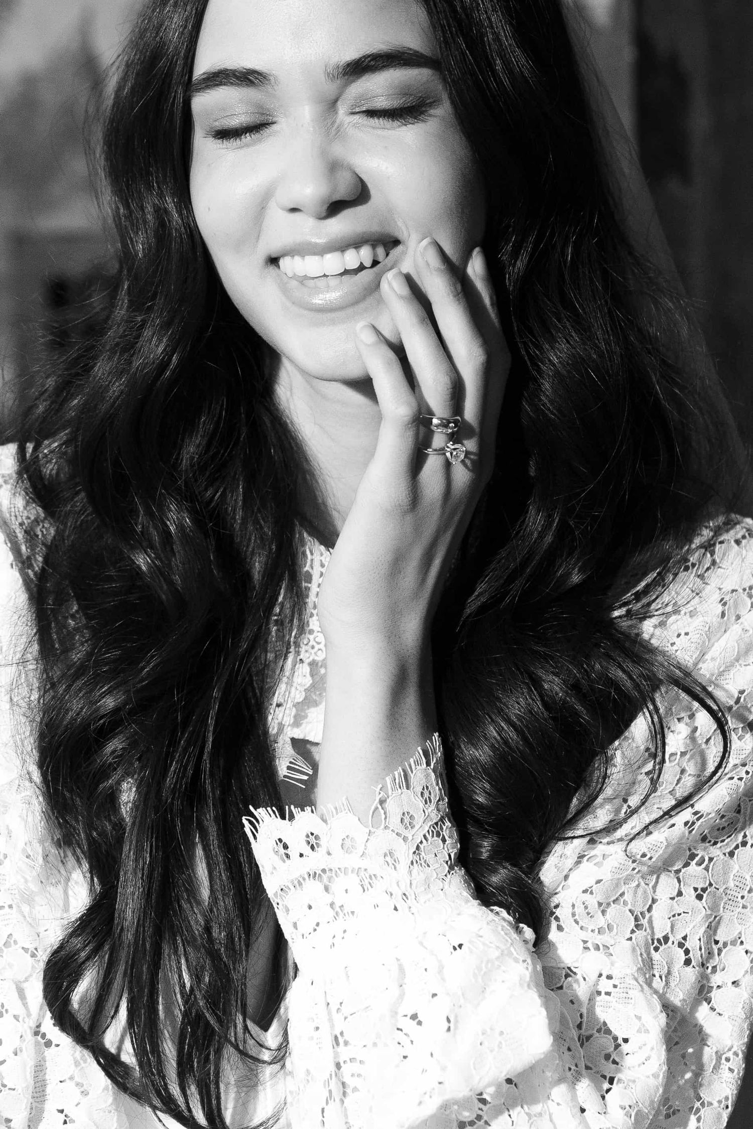 Black and white portrait of a smiling young woman with long wavy hair, wearing a lace top and ring.