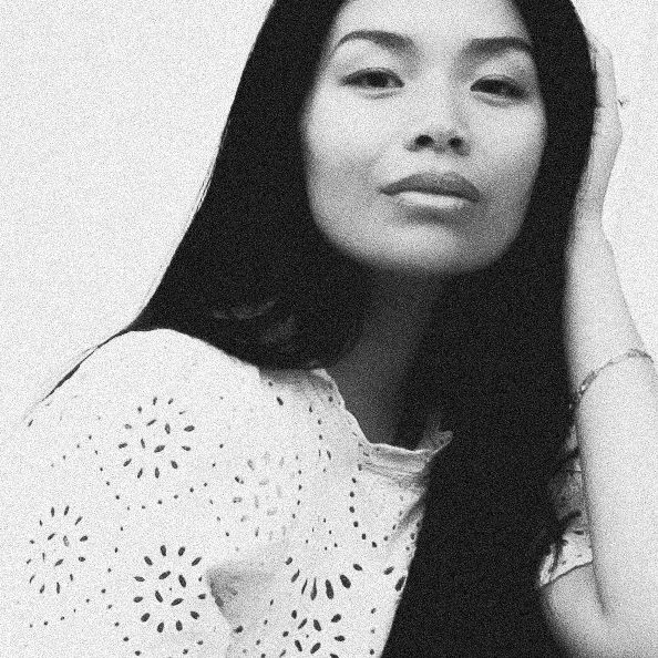 Black and white portrait of a young woman with long dark hair wearing a white eyelet top, hand raised near her face.