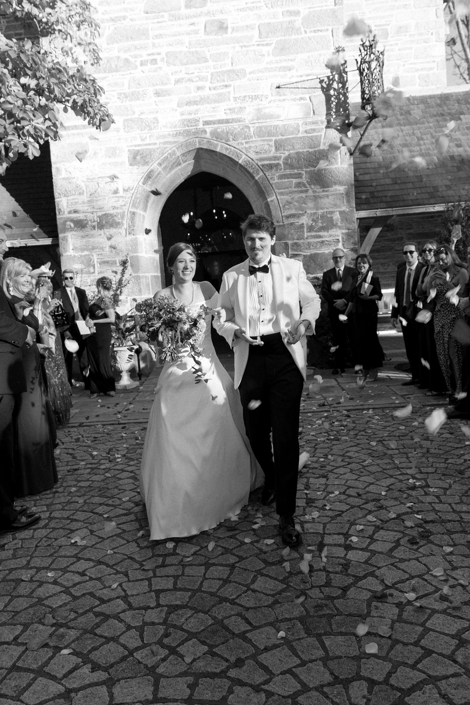 Black and white photo of a bride and groom exiting a stone church as guests celebrate with confetti.
