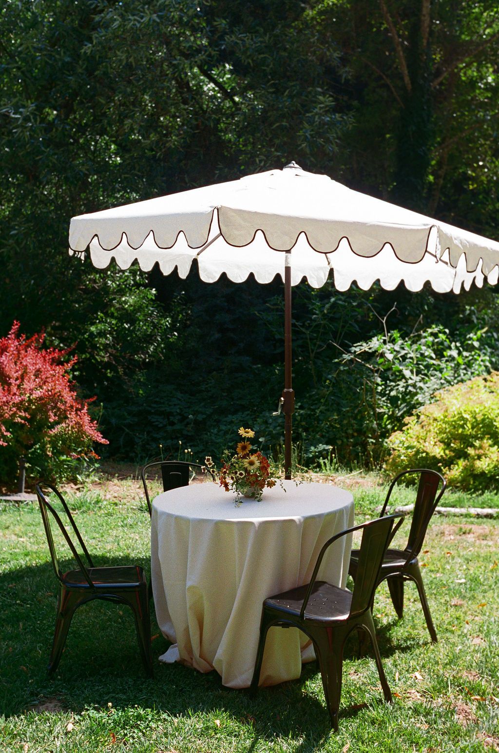 White umbrellas and round tables set up for an outdoor garden reception under tall trees.
