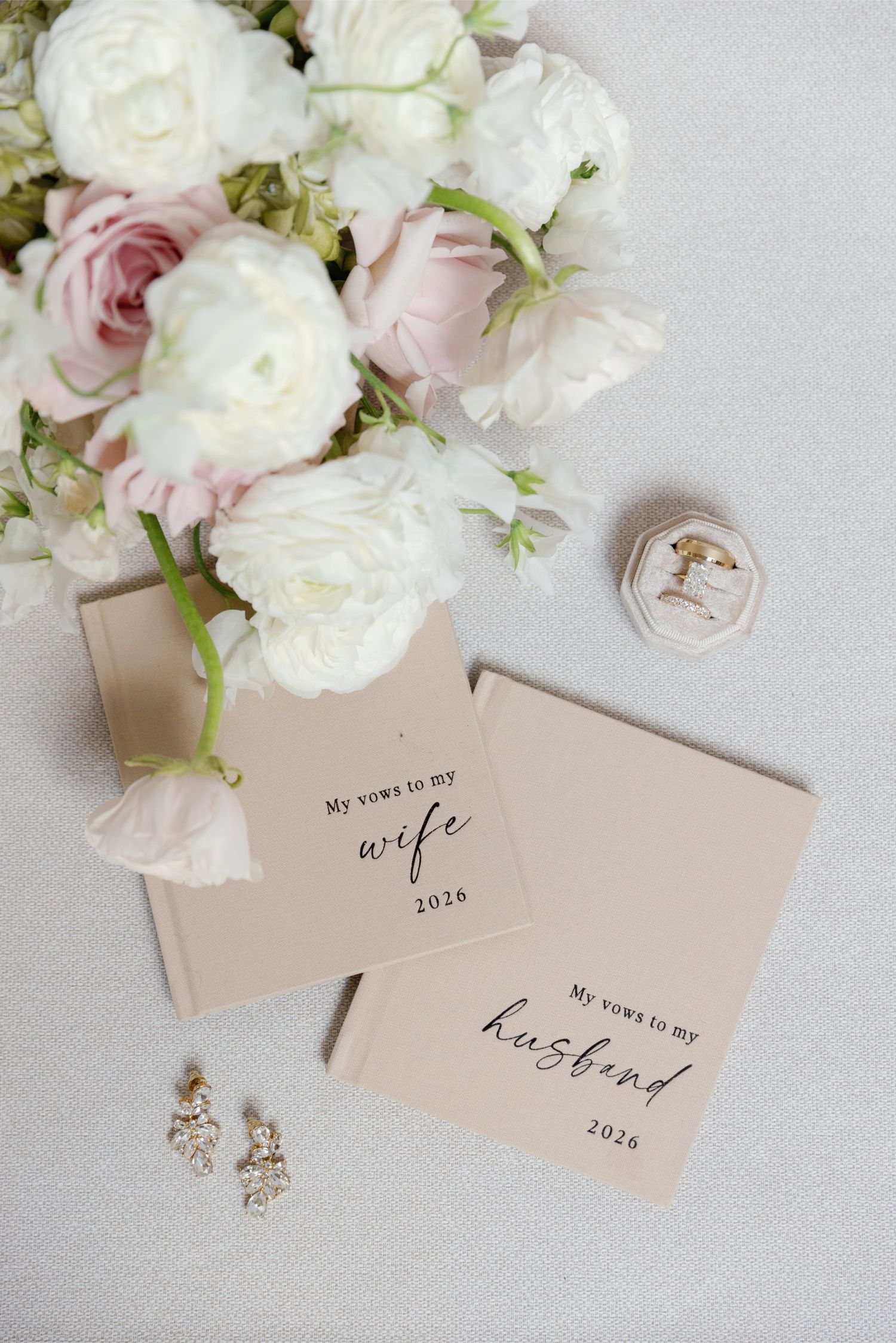Elegant wedding vow books for husband and wife, styled with blush roses, crystal earrings, and a ring box on a neutral backdrop.